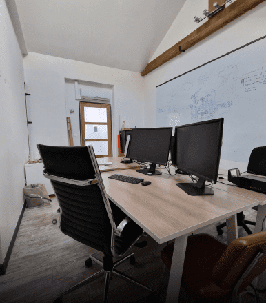 Image de la salle de coworking. Sur l'image figurent 2 écrans d'ordinateur au centre d'une grande table rectangulaire. Différents sièges sont disposés autour de la table. En arrière plan il y a un grand tableau blanc sur lequel sont inscrit différentes réflexions. Elle ne sont pas lisible sur l'image.