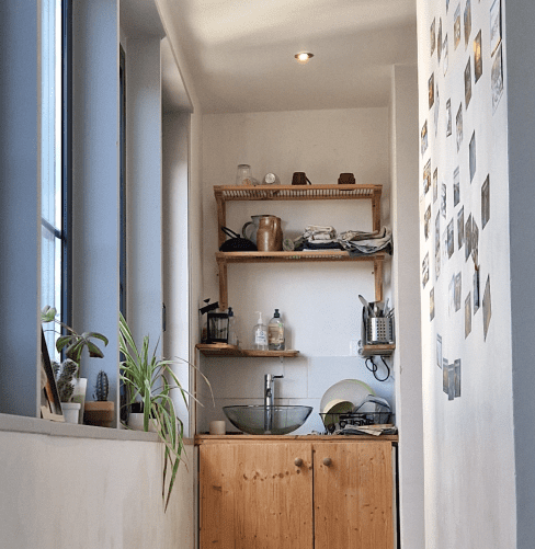 Image de la kitchenette de l'Escale&Co. Située au fond d'un couloir lumineux, la kitchenette est composée d'une vase transparente, d'un égouttoir à vaisselle et d'étagères sur lesquelles sont rangés différents ustensiles de cuisine (couverts, bouilloire, liquide vaisselle, torchons, tasses)